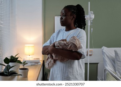 Woman cradling newborn baby in hospital room with a bedside table and plants surrounding. IV stand and a cozy lamp illuminating the background - Powered by Shutterstock