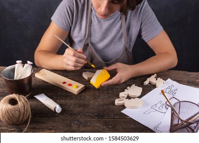  A woman creates a Christmas tree toy in her workshop. Paints a wooden blank in yellow. Gray background. - Shutterstock ID 1584503629