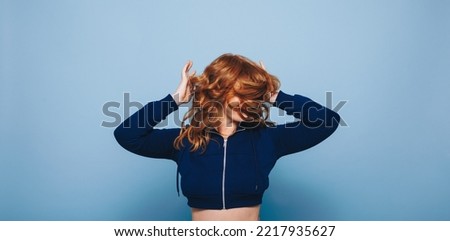 Woman in a crop top dancing and flipping her ginger hair excitedly. Happy young woman celebrating and having fun while standing against a blue studio background.