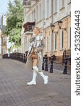 A woman crossing a cobblestone street in a fashionable autumn outfit, holding a small handbag and wearing knee-high boots amid lovely architecture.