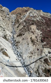 Woman Crossing A Dangerous Suspension Bridge, Along The Landslide Area On The Way To Tilicho Base Camp, Annapurna Circus, Himalayas, Nepal. Dry And Desolated Landscape. Steep And Sharp Slopes. Danger