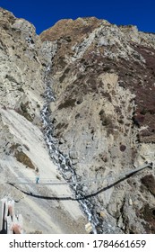 Woman Crossing A Dangerous Suspension Bridge, Along The Landslide Area On The Way To Tilicho Base Camp, Annapurna Circus, Himalayas, Nepal. Dry And Desolated Landscape. Steep And Sharp Slopes. Danger