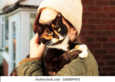A woman cuddles (tortoiseshell) a calico cat against her face outside a suburban home.