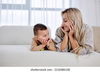 Woman Cuddling Talking To Small Kid Son, Having Fun Together On Sofa In Studio Living Room. Smiling Child Boy With Blonde Woman Mother, Laughing At Home, Look At Each Other. Leisure Funny Time