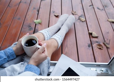 Woman with cup of coffee sitting relaxing home in the window, she's covered in a blanket. Fall leaves on wooden patio deck. Autumn winter concept