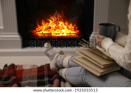 Woman with cup of hot drink and book resting near fireplace at home, closeup