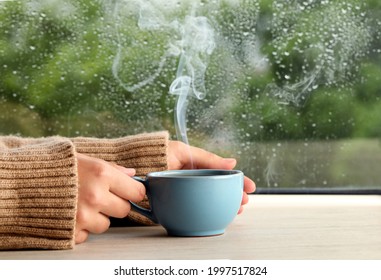 Woman with cup of hot drink at wooden table near window on rainy day, closeup - Powered by Shutterstock