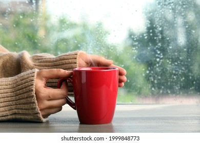 Woman With Cup Of Hot Drink At Wooden Table Near Window On Rainy Day, Closeup