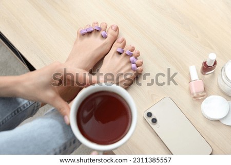 Woman with cup of tea preparing toenails for pedicure on table at home, closeup