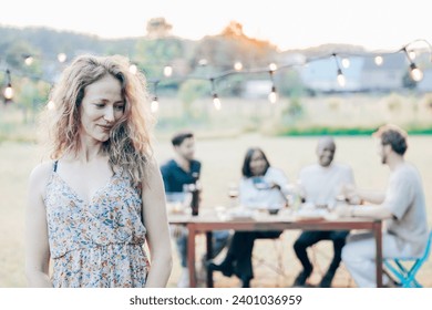 A woman with curly blonde hair, dressed in a floral dress, stands thoughtfully in the foreground, a soft smile playing on her lips. Behind her, a group of friends enjoys a meal outdoors, their - Powered by Shutterstock