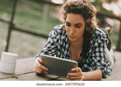 Woman with curly braided hair, wearing a checkered shirt, lies on her stomach, engrossed in a tablet. Her focused expression highlights her deep concentration, with a coffee mug beside her - Powered by Shutterstock