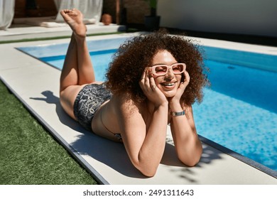 Woman with curly hair, bikini, pink sunglasses, smiling by pool edge, relaxed and happy - Powered by Shutterstock