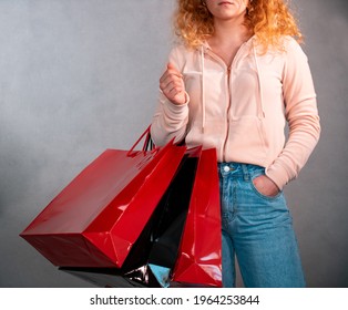 Woman with curly hair, blue jeans and rose hoodie carries shopping bags in black and red. 