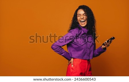 Woman with curly hair enjoys using her smartphone against a vibrant orange background. She looks happy and casual, adding a touch of fashion and excitement to the scene.