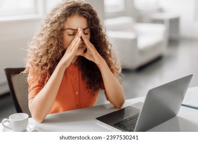 Woman with curly hair feels stress, rubbing the bridge of her nose with eyes closed, sitting at a laptop in an office, with a cup of coffee beside her - Powered by Shutterstock