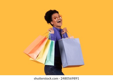 A woman with curly hair and heart-shaped sunglasses is smiling widely and holding several colorful shopping bags. She is standing against yellow background, suggesting a joyful shopping experience. - Powered by Shutterstock