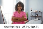 Woman with curly hair and pink uniform standing with arms crossed in a spa room, indicating wellness and beauty services provided by a smiling professional in an indoor salon setting