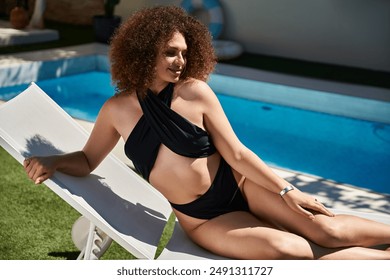 A woman with curly hair relaxes in a black swimsuit on a lounge chair near a pool. - Powered by Shutterstock