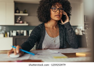 Woman With Curly Hair Using Desktop Computer And Talking On Cell Phone. Female Architect Working At Her Desk At Home Office.