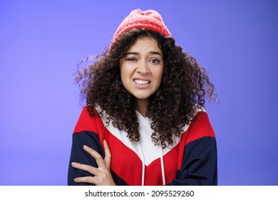 Woman with curly hair in winter beanie feeling uncomfortable and discomfort clenching teeth and frowning intense as hugging herself insecure and awkward, unwilling to say cruel rejection, feel awkward