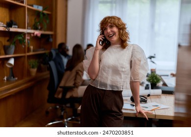 A woman with curly red hair is smiling while talking on the phone in a modern office setting. She is leaning against a desk with papers and a plant on it. - Powered by Shutterstock