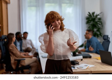 A woman with curly red hair is smiling while talking on the phone in a modern office setting. She is leaning against a desk with papers and a plant on it. - Powered by Shutterstock