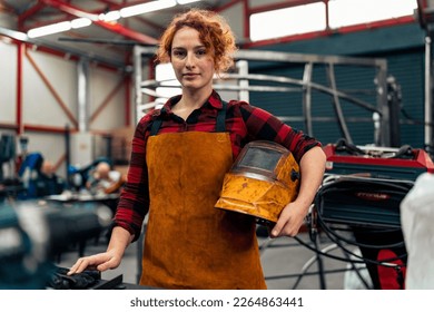 Woman with curly red hair standing in workshop, holding protective equipment under arm and smiling at camera, workshop in background - Powered by Shutterstock