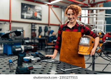 Woman with curly red hair standing in workshop, holding protective equipment under arm and smiling at camera, workshop in background - Powered by Shutterstock