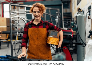 Woman with curly red hair standing in workshop, holding protective equipment under her arm and smiling at camera, workshop in background - Powered by Shutterstock