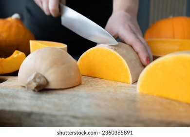 Woman cutting butternut squash pumpkin into slices with a kitchen knife on a wooden board. Fresh raw sliced pumpkins pieces. Preparing ingredients for a seasonal autumn fall dish, soup or pie.