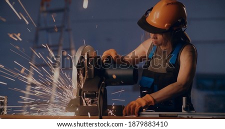 Woman cutting metal pipe with electric miter saw and measuring diameter with calipers working in dark factory workshop