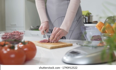 Woman Cutting Raw Meat On Small Pieces With Kitchen Knife Cutting Board. Woman Puts Pieces Of Raw Meat In A Glass Bowl. Female Cutting Red Beef Filet On Wooden Board, Holding Knife In Hand.