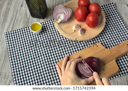 Woman cutting a red onion on a wooden board, opposite another wooden board with tomatos, garlic and half red onion, next to botlle olive oil and small bowl with olive oil