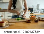 A woman is cutting vegetables on a wooden cutting board. She is wearing a white shirt and blue jeans. The vegetables include carrots, celery, and lettuce
