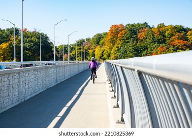 A Woman Cycling On A Bridge Over Credit River On A Sunny Autumn Day With Colorful Foliage In The Background