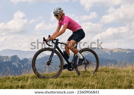 Woman cyclist in cycling kit and helmet riding a gravel bicycle on mountain road.Sportswoman cycling in the beautiful natural surroundings. Cyclist practicing on gravel road.Bucegi Mountains, Romania.