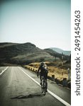 A woman cyclist training on a lonely road on a sunny day wearing a black jersey and bib shorts and a white helmet. 