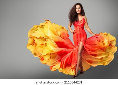 Woman Dancing in Colorful Yellow Orange Flying Dress. Happy Girl Model in Creative Bright Gown over Gray Background. Curly Latin Salsa Dancer