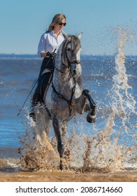 Woman and dapple gray Spanish horse. Andalusian horse stomps his hoof on sea and splashes water. Summertime vertical outdoors image.