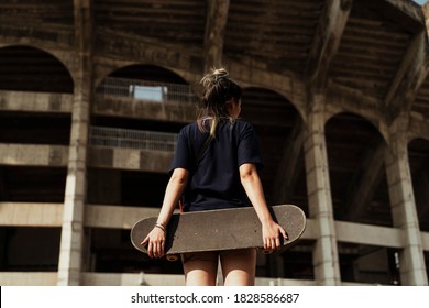 Woman In Dark Blue Polo Shirt Hold The Longboard At Her Butt Standing In Front Of Pretended Colosseum Building.