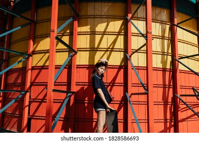 Woman In Dark Blue Polo Shirt Standing In Front Of Yellow And Red Wall In Summer Sunlight With Longboard.