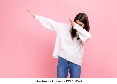 Woman with dark hair showing popular internet meme pose, celebrating success victory, dabbing trends, wearing white casual style sweater. Indoor studio shot isolated on pink background.