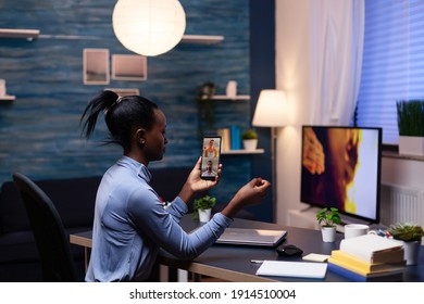 Woman With Dark Skin Holding Phone In Online Meeting Sitting At Desk In Hime Office Late At Night. Busy Employee Using Modern Technology Network Wireless Doing Overtime For Job.