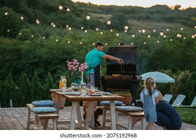 Woman And Daughter Is Playing With Dog After They Arranged Dining Table In The Backyard. Young Adult Man Barbecuing Beside Them