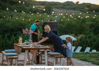 Woman and daughter setting up dining table in the backyard for family gathering while man barbecuing beside