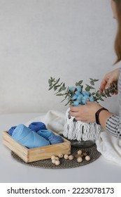 A Woman Decorates A Christmas Table Arrangement. On A White Table, A Wooden Box With Blue Color Balls Of Threads, A Knitted Stand And A Jar, Lined With Macrame