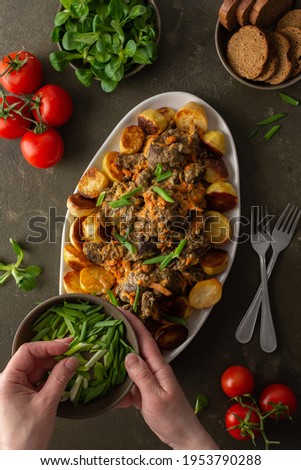 Woman decorates with green onions a dish with stewed wild goat and baked potatoes, Mache salad, bread and tomatoes on a dark background, top view