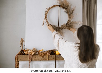 Woman decorating fireplace with stylish autumn wreath. Rustic wreath with dried grass, herbs and ribbon in hands. Fall decor and arrangement in home. Thanksgiving and Halloween - Powered by Shutterstock