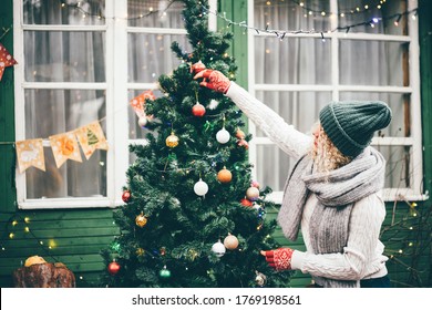 Woman Decorating Home For Christmas And Hanging Baubles On Christmas Tree Outside. 