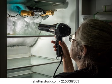 Woman defrosting the inside of a fridge where there are some bottles, with the help of a hair dryer.